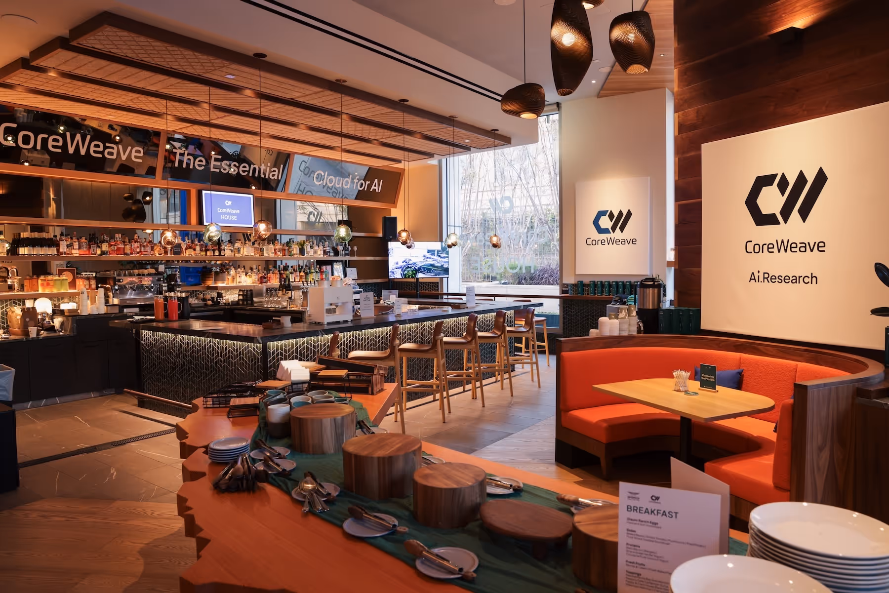 Modern cafe interior with wooden bar stools, orange cushioned booth seating, and CoreWeave AI research branding on the walls.