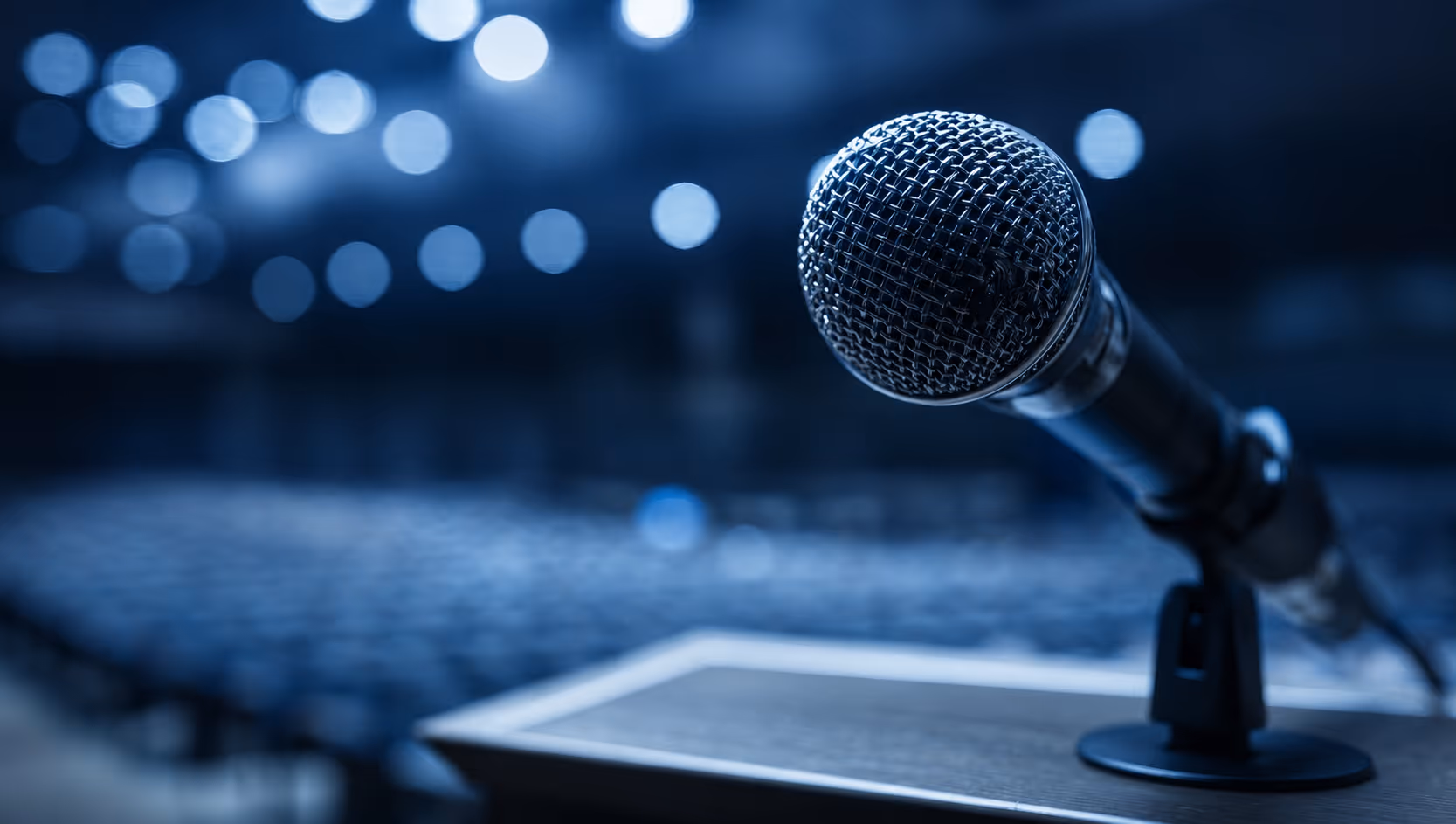 Close-up of a microphone on a stand with blurred auditorium and lights in the background.
