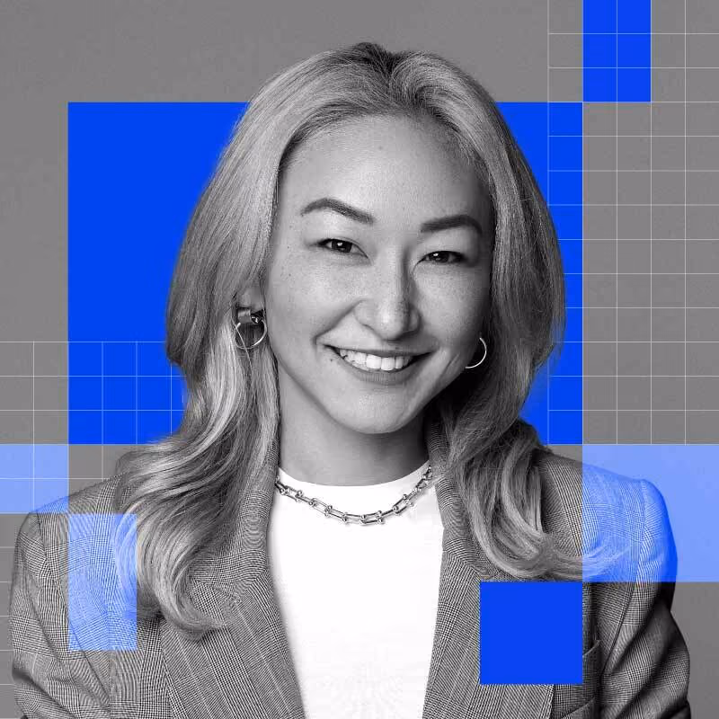 Smiling woman with long hair wearing a suit jacket and chain necklace against a gray and blue background.