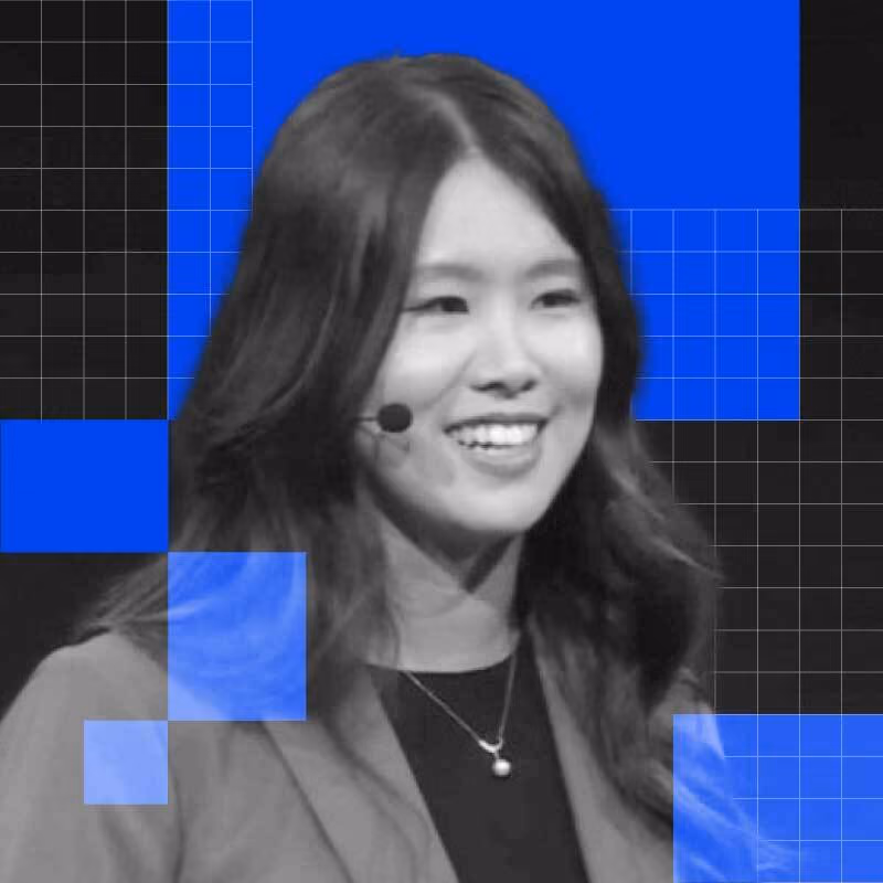 Smiling woman with long dark hair wearing a microphone and necklace on a black background with blue squares.