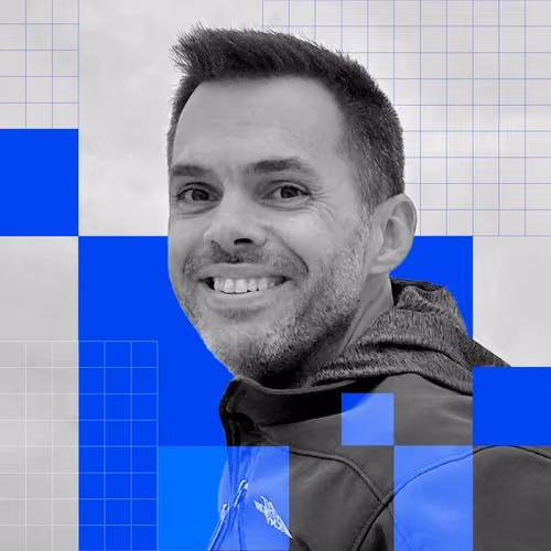 Smiling man with short hair and light beard wearing a jacket against a gray and blue geometric background.