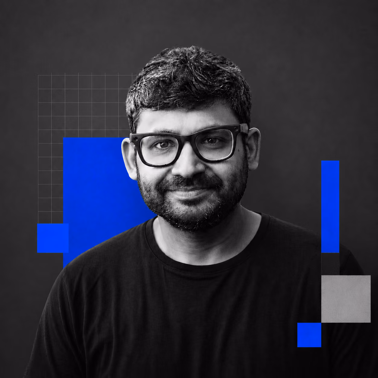 Smiling man with glasses and a beard wearing a black shirt on a dark background with blue and gray shapes.