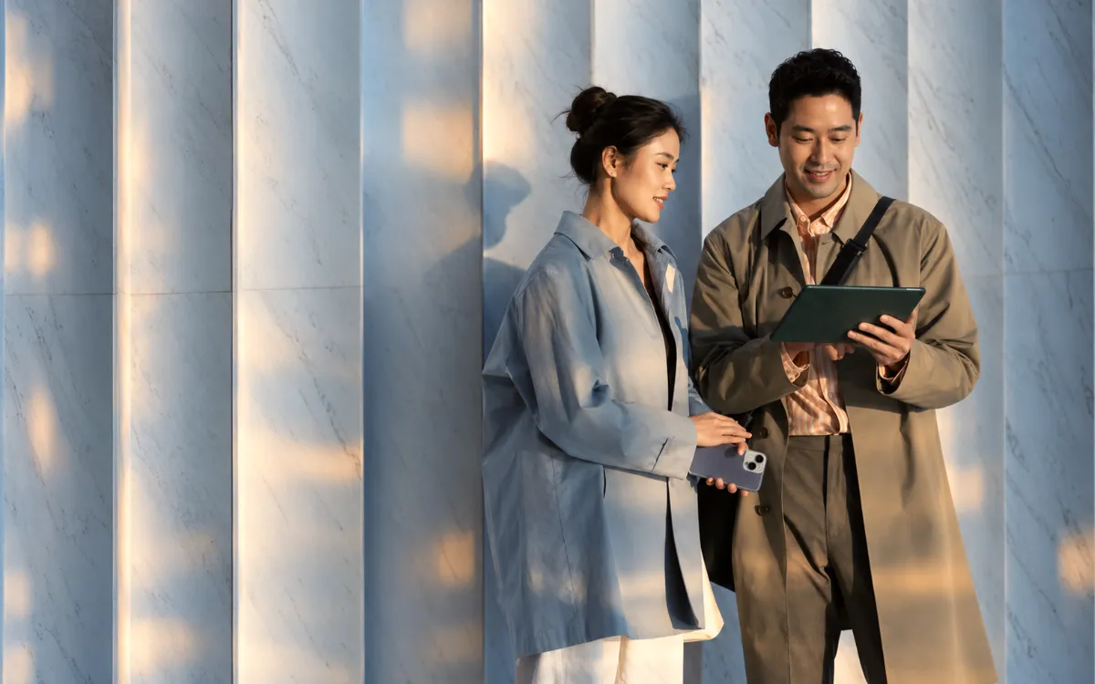 Two people standing in front of a marble wall with vertical panels, one holding a tablet and the other holding a smartphone, both looking at the tablet and smiling.