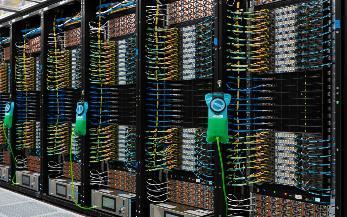 Row of server racks filled with numerous network cables in blue, yellow, and white, connected to servers and hardware components with green cooling or monitoring devices attached to some racks.