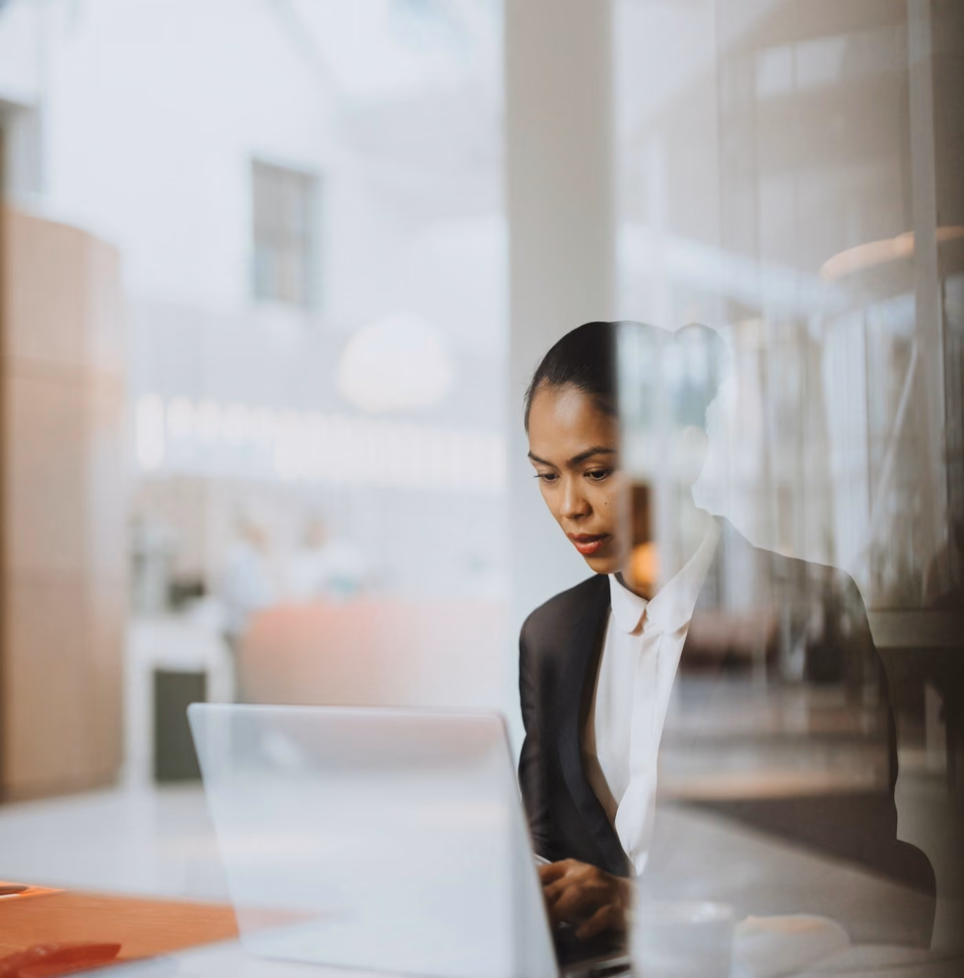 A focused woman in business attire working on a laptop in a modern office with soft natural light and blurred background.