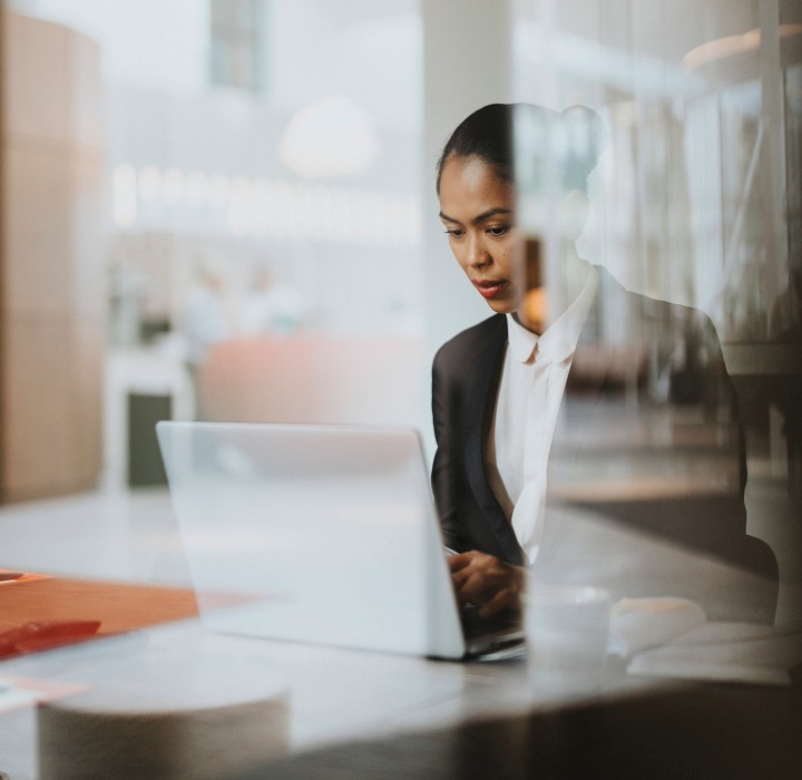A woman in business attire sits indoors at a table working on a laptop, looking focused.