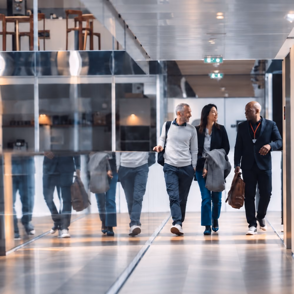 Three professionals walking down a modern office hallway, engaged in conversation, with glass walls reflecting their images.