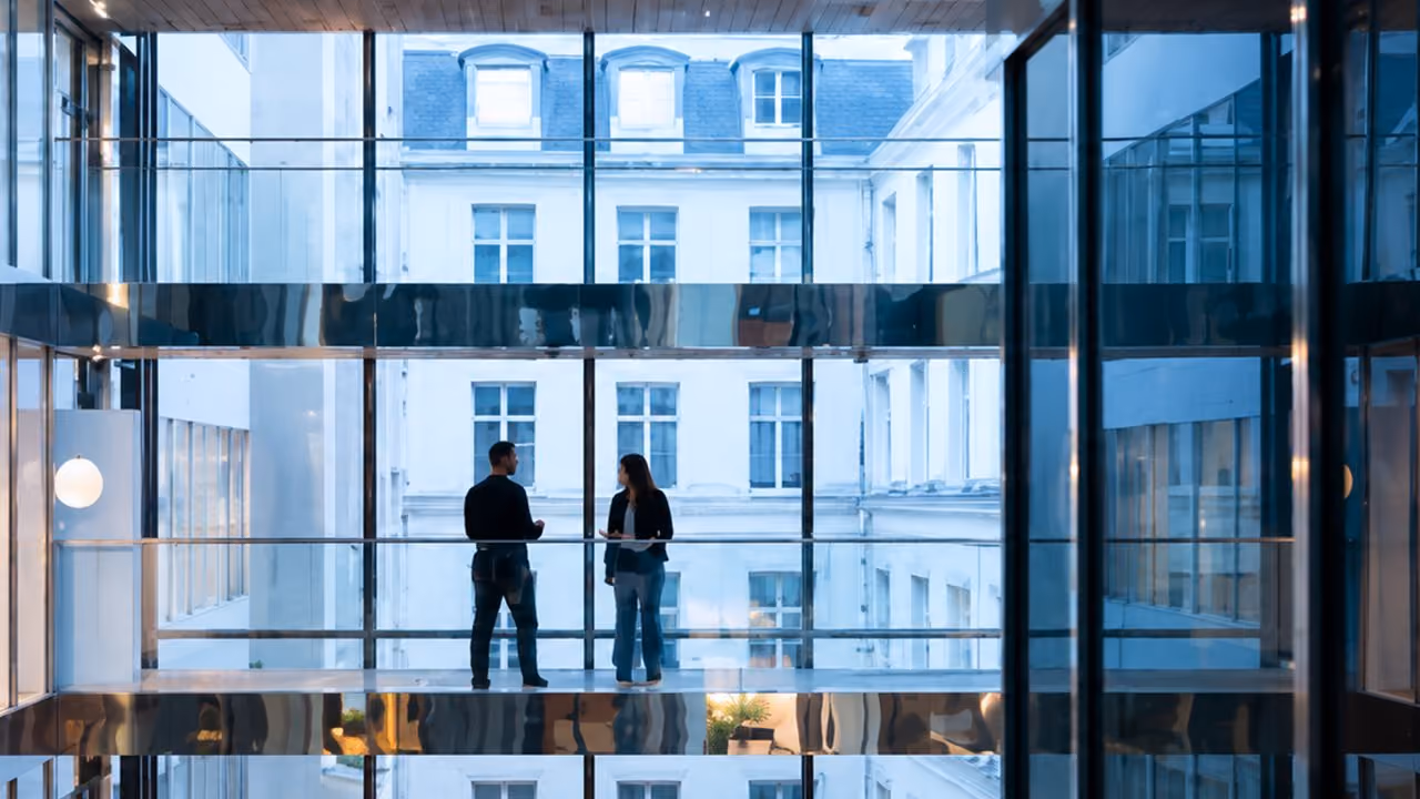 Two people standing opposite each other on a glass-walled walkway inside a modern building with large windows showing an exterior view of a classic architectural facade.