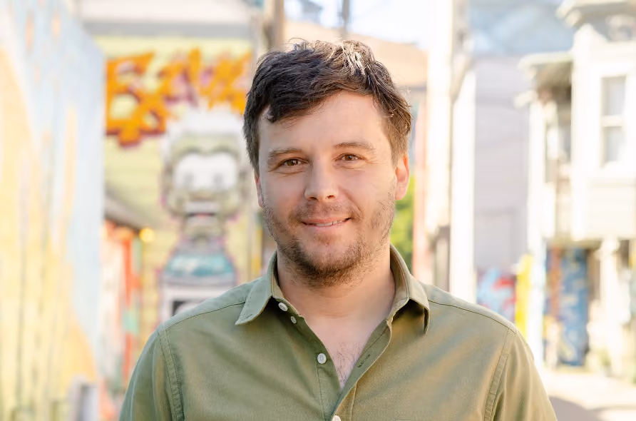 Man with short brown hair and light beard wearing an olive green button-up shirt, standing outdoors with graffiti-covered walls and buildings blurred in the background.