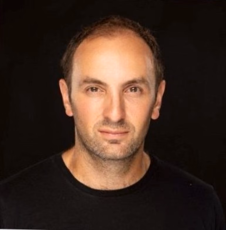 Portrait of a man with short dark hair and light stubble wearing a black crew neck shirt against a black background.