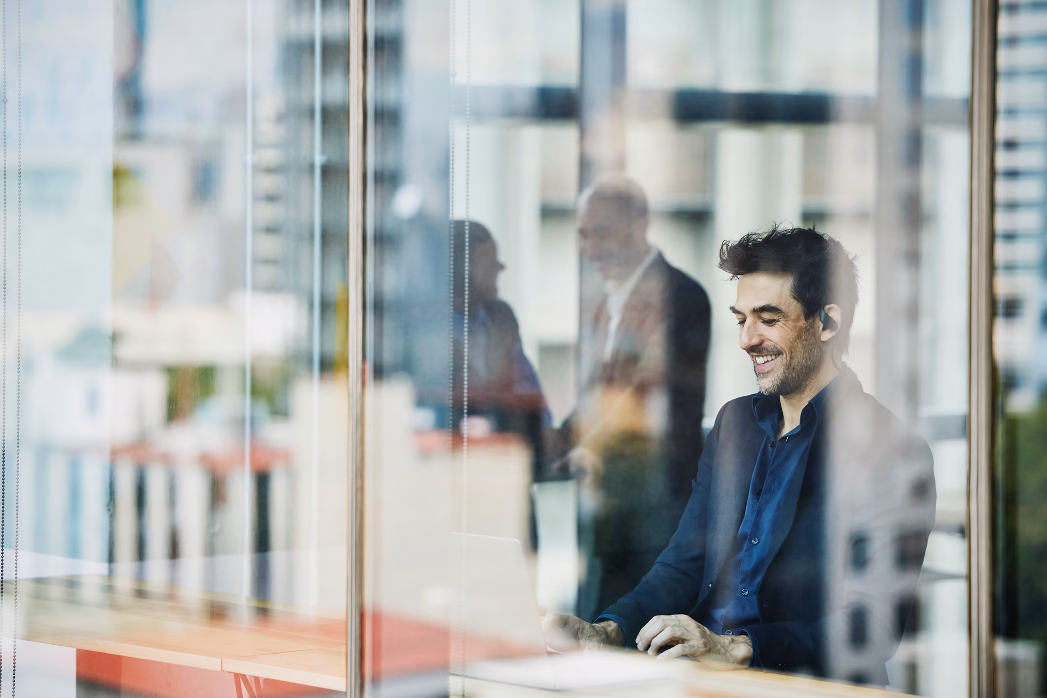 Man in a dark suit sitting at a desk, smiling while working on a laptop in an office with glass walls, with reflections of two people shaking hands in the background.