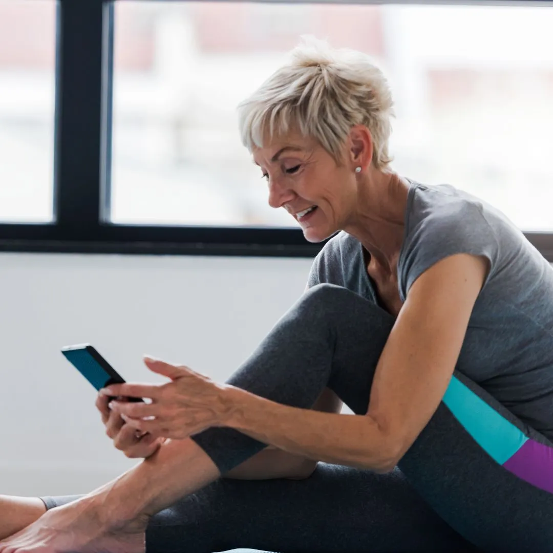 Older woman sitting indoors in athletic wear, smiling as she looks at a smartphone.
