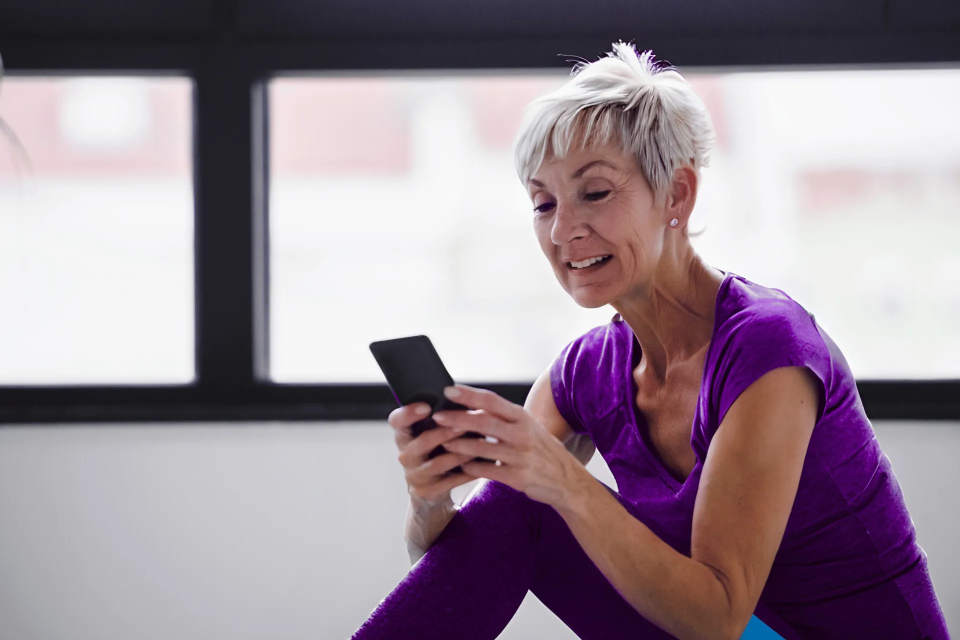 Woman in purple athletic wear sitting and smiling while using a smartphone indoors.