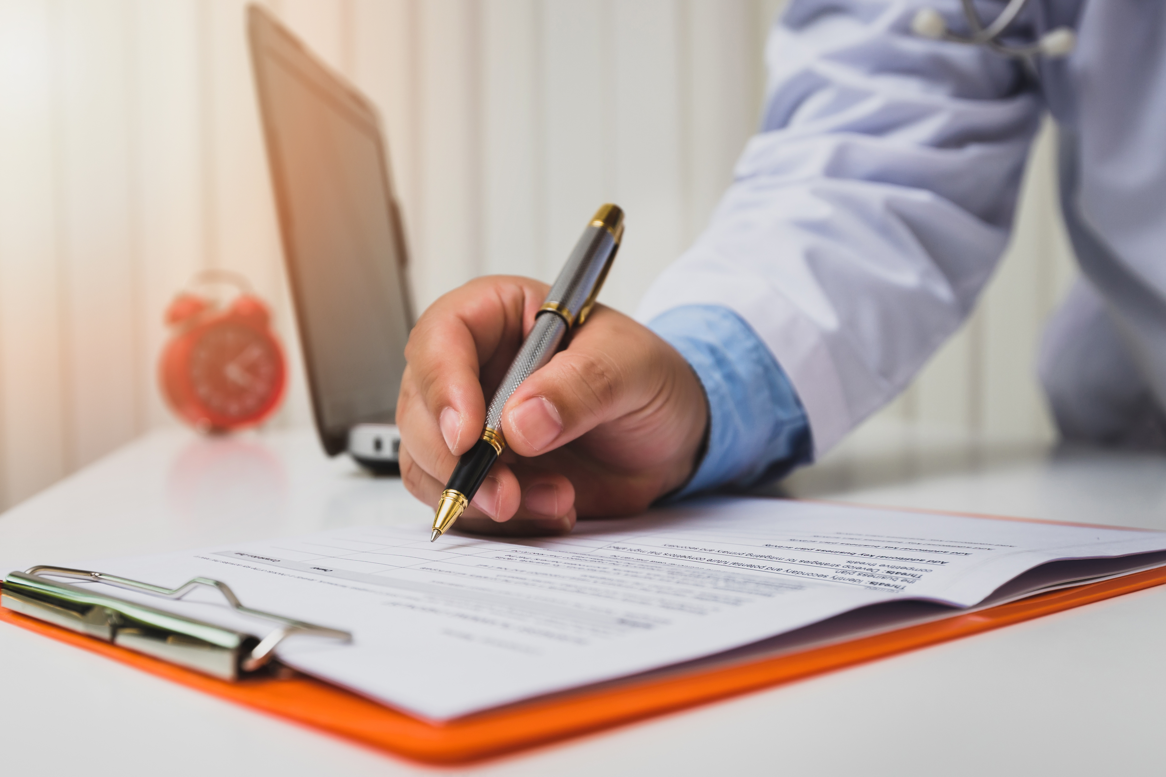 A person in a white coat writes on a document attached to a clipboard, with a laptop and alarm clock on the desk.