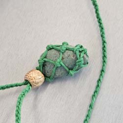 Green stone wrapped in green macrame cord with a small beige bead on a gray surface.
