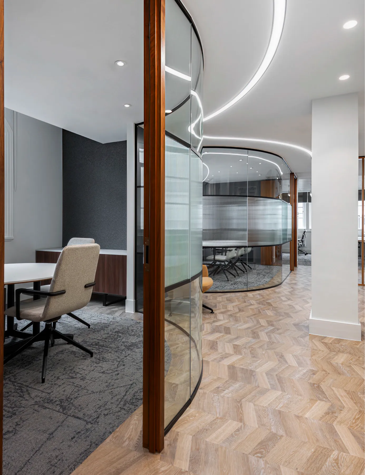 Office corridor with wood flooring, curved ceiling lights, and glass meeting room walls.