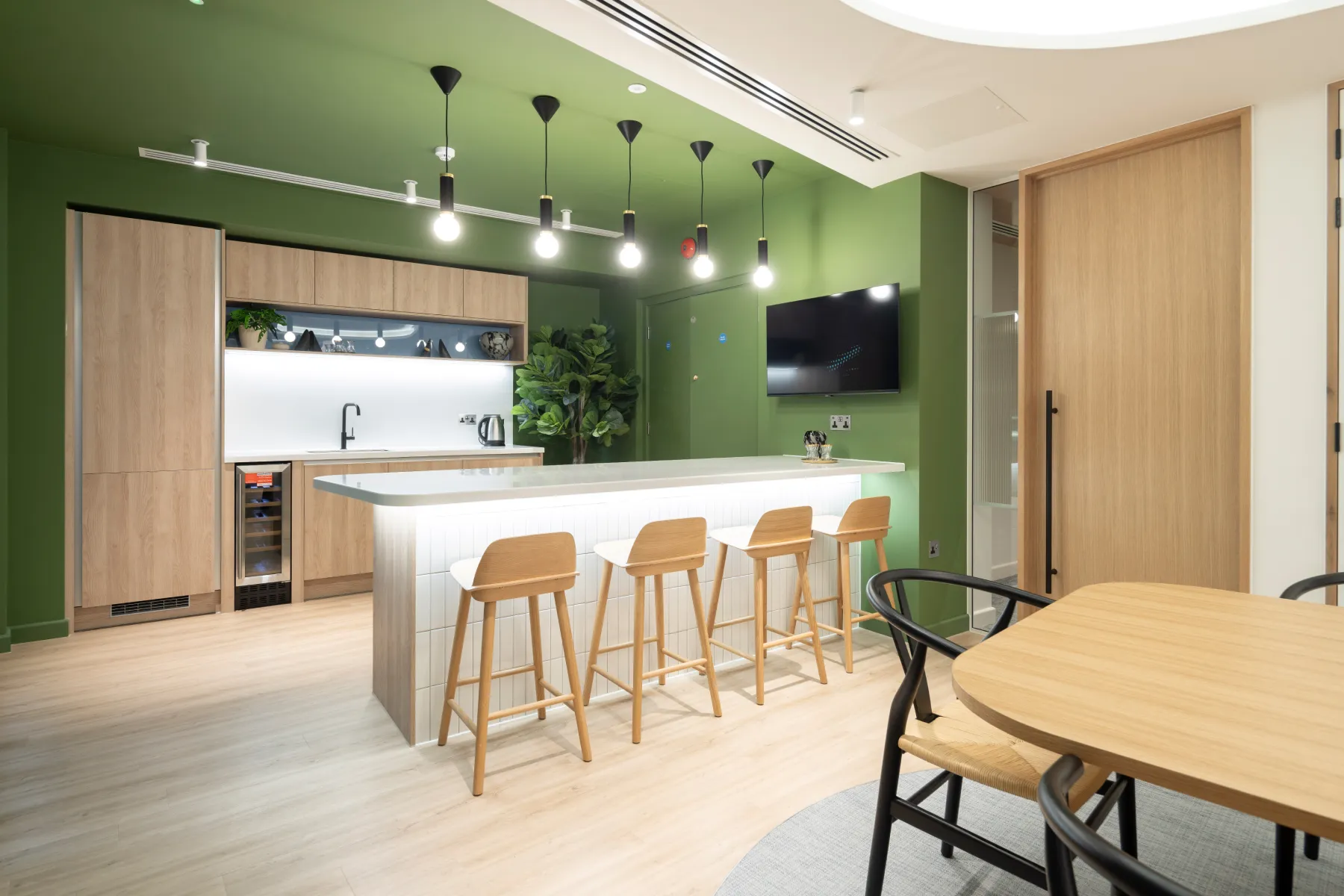 Contemporary office kitchen area with green walls, wooden cabinetry, and high stools at a breakfast bar.