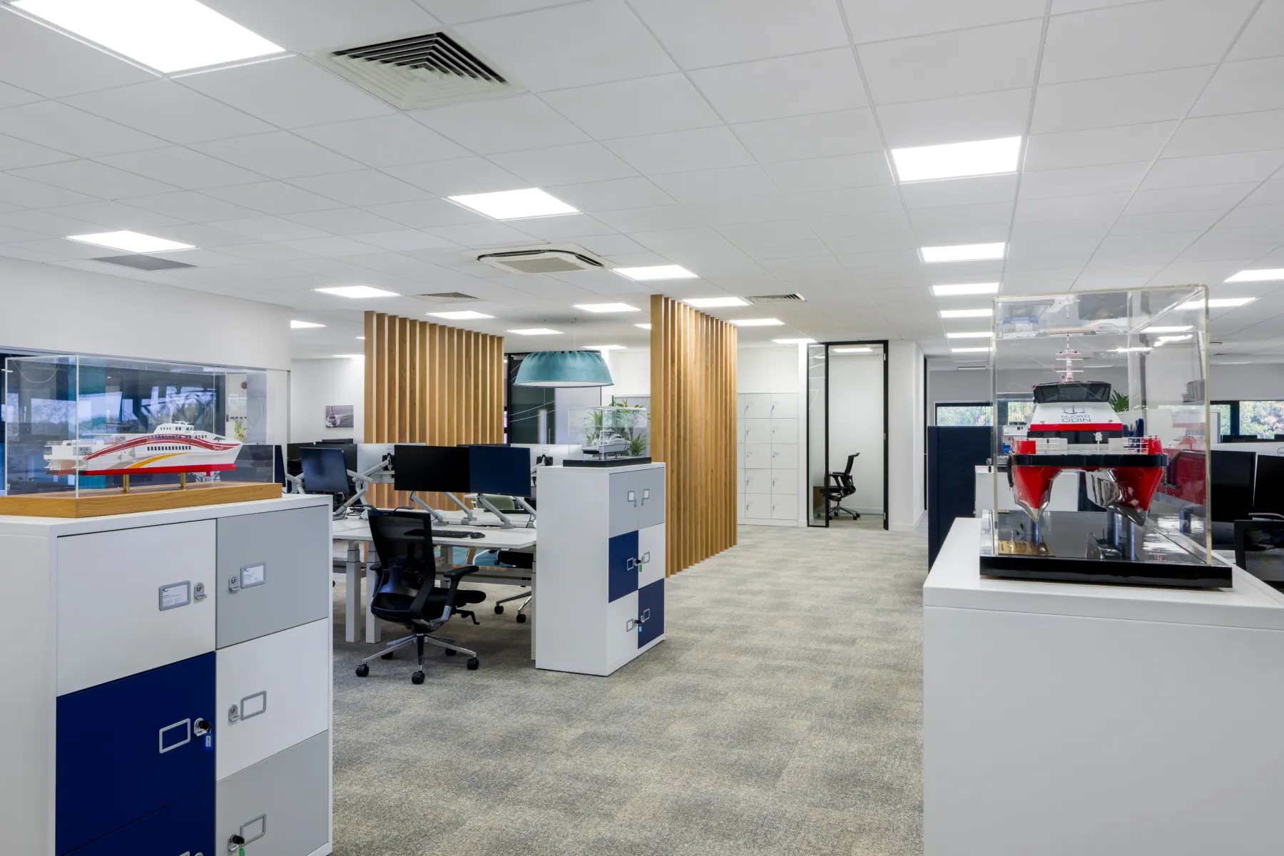 Open-plan office featuring height-adjustable desks, wooden slatted dividers, office storage, and model vessels displayed in acrylic cases.