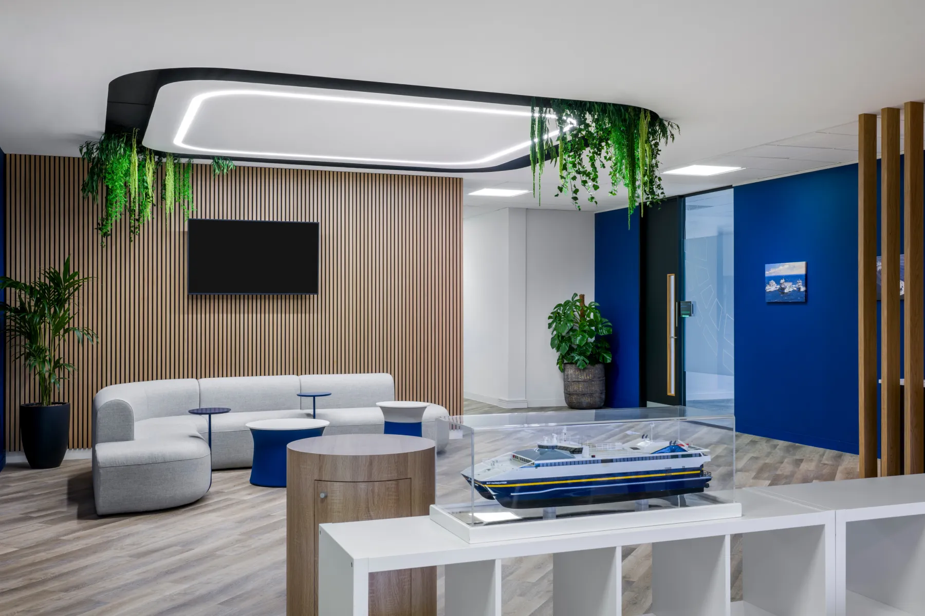 Modern reception lounge with curved grey seating, blue side tables, timber slat walling, and suspended planting framing a feature light.