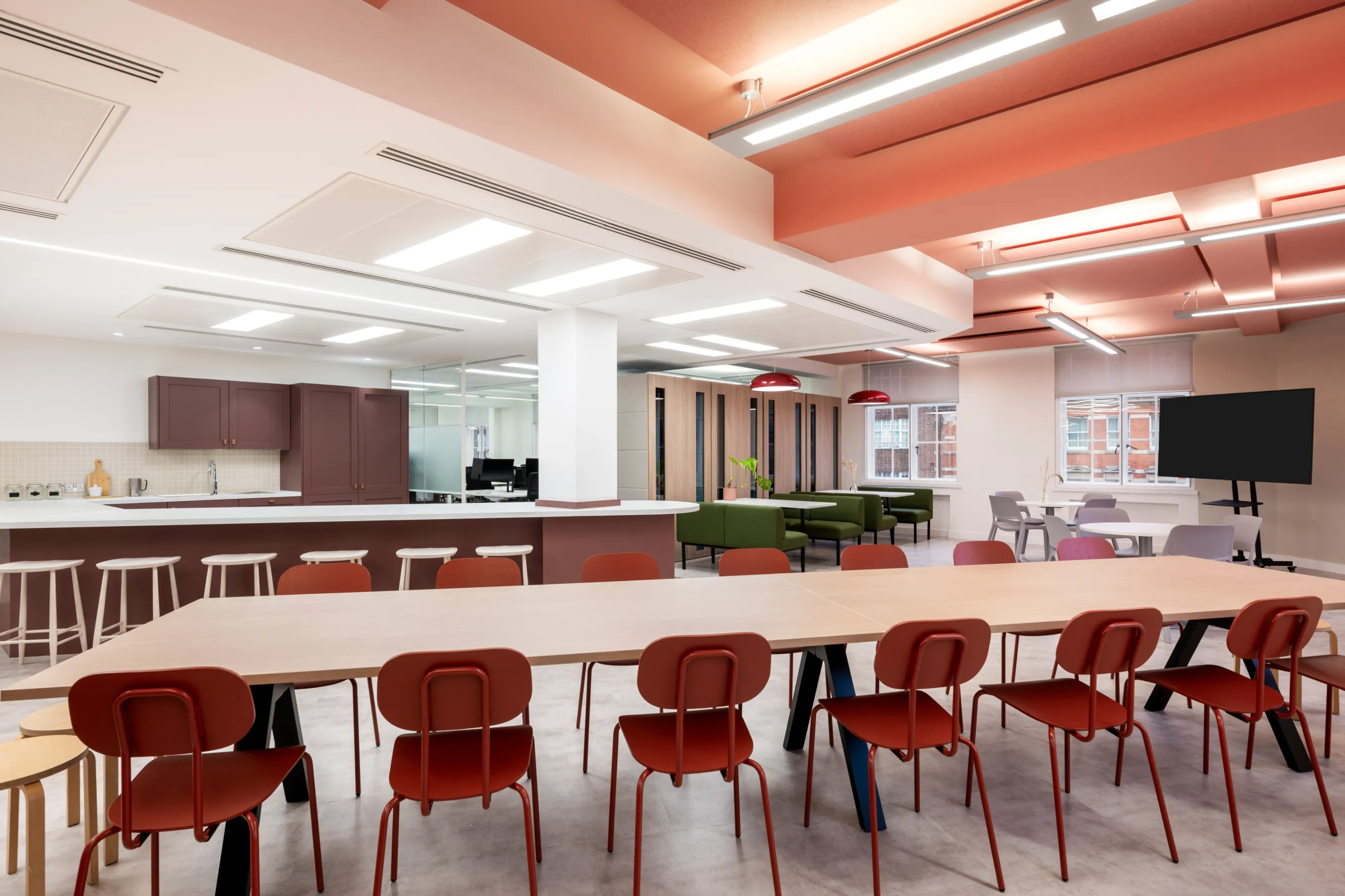 Bright office breakout area with long desks, red chairs, and exposed ceiling details.