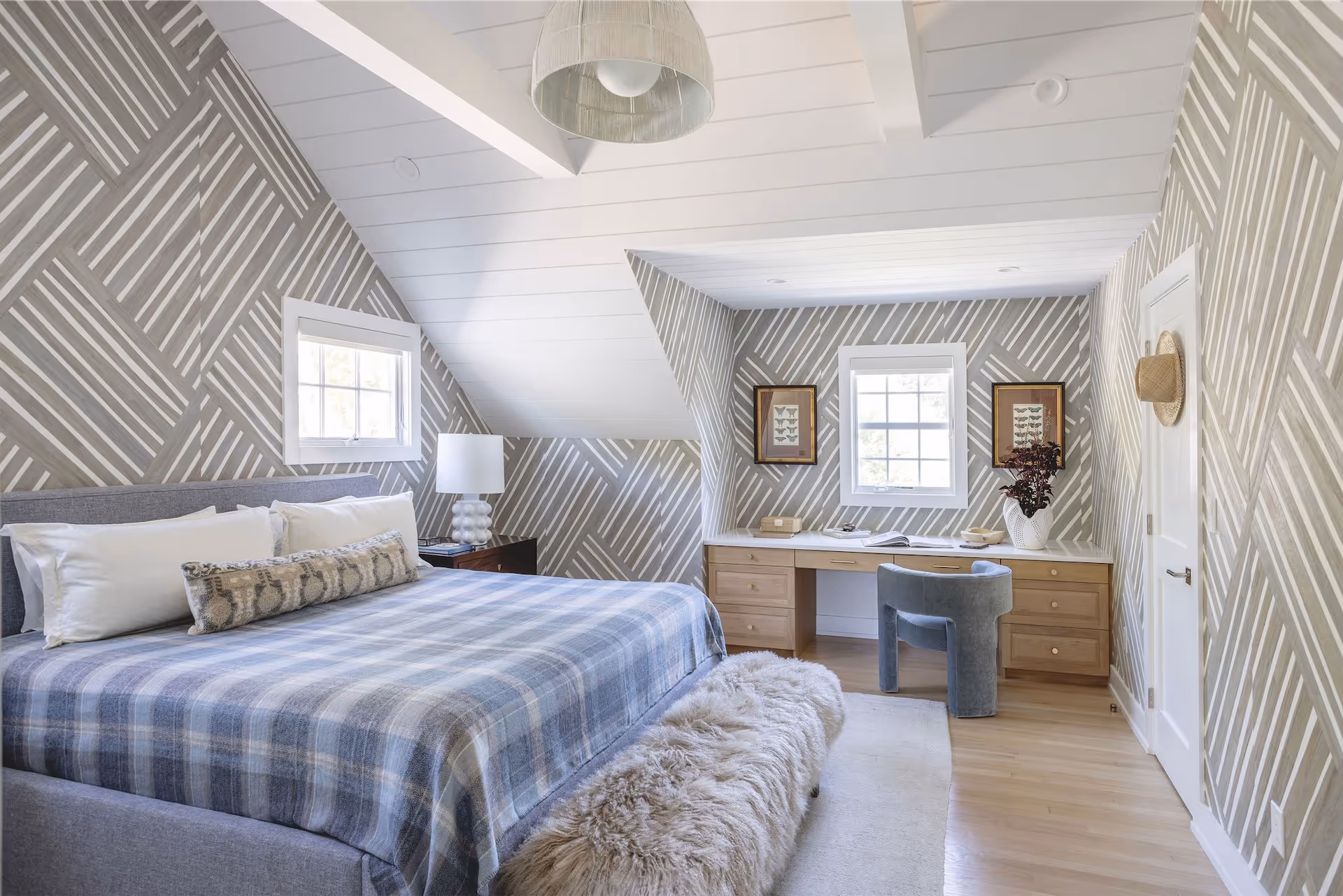 Modern attic bedroom with geometric striped wallpaper, a gray bed with plaid blanket, wooden desk under window, and a blue velvet chair.