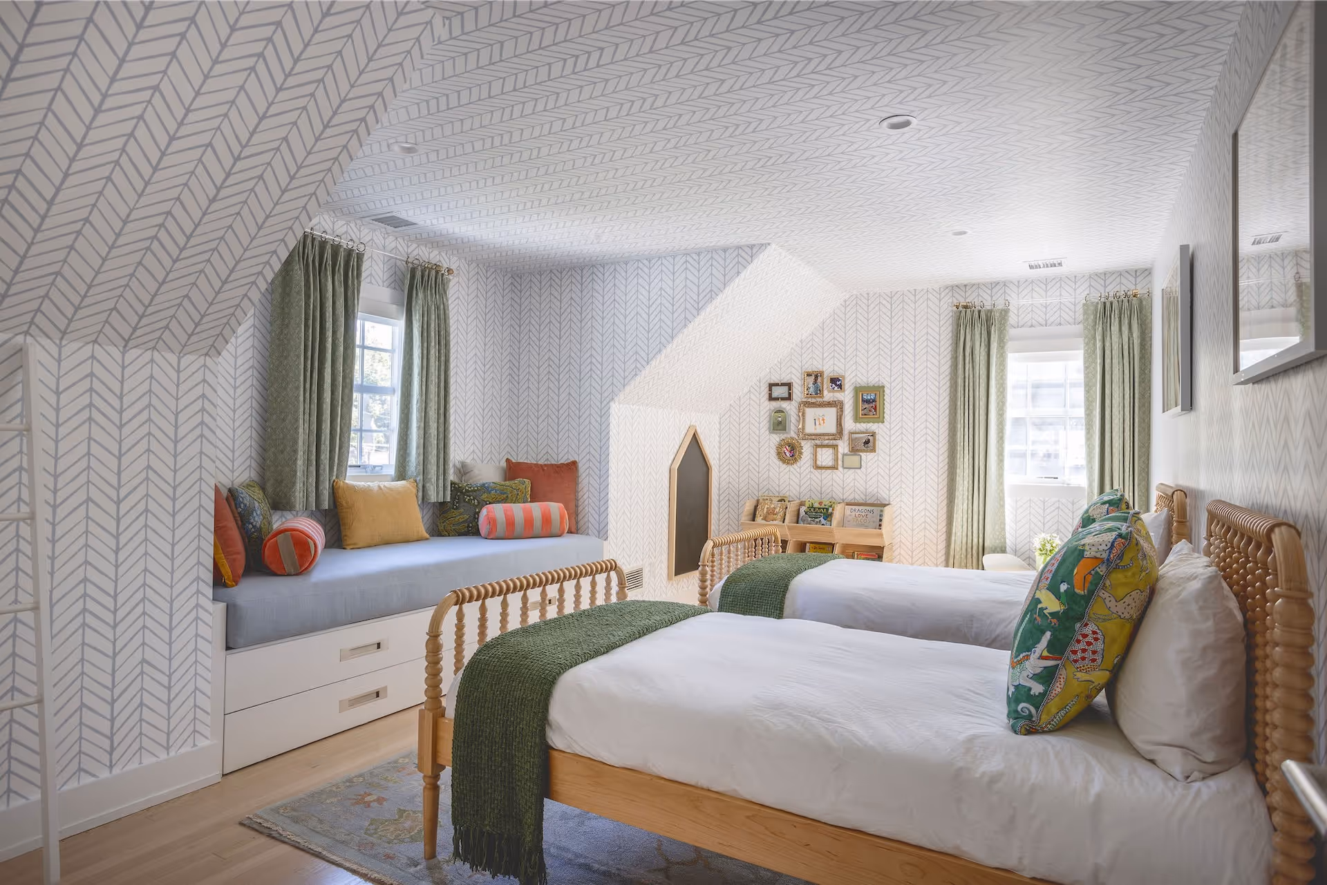 Bright kids' bedroom with two wooden twin beds with white bedding and green throws, a window seat with colorful pillows, and patterned wallpaper throughout.