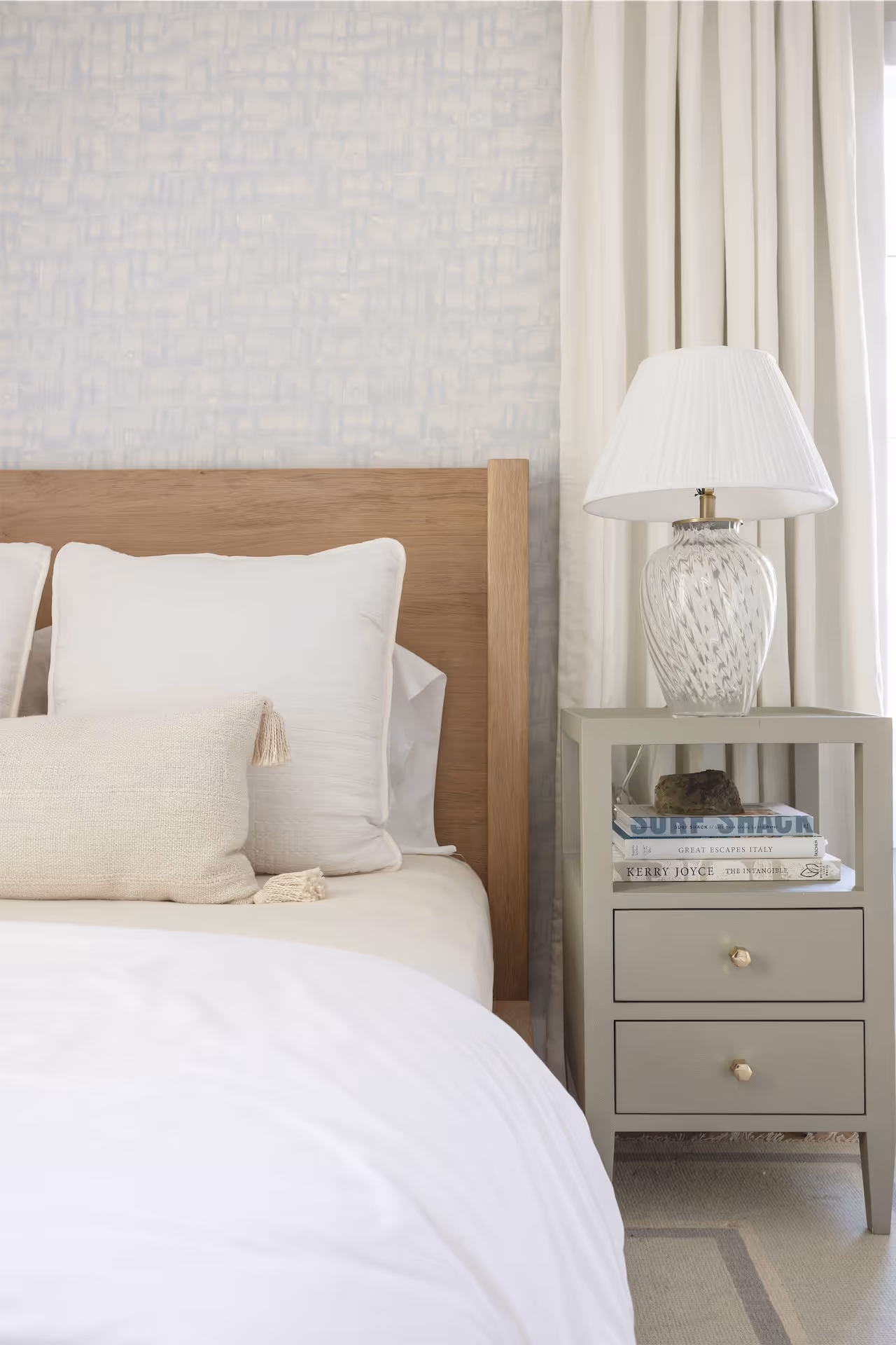 Neutral bedroom with a light wooden headboard, white pillows, a beige textured lumbar pillow, and a gray nightstand holding a glass lamp and stacked books.