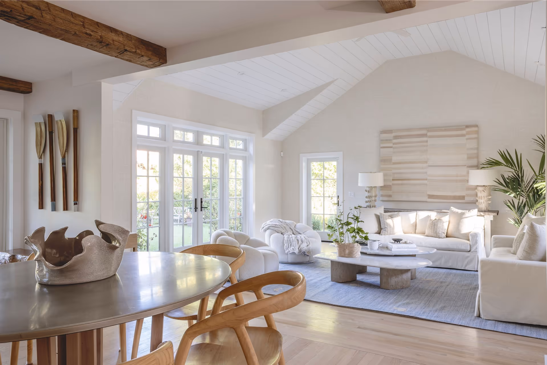 Bright modern coastal living room with wooden ceiling beams, white sofas, round coffee table, and stylish wooden dining chairs around a round table.