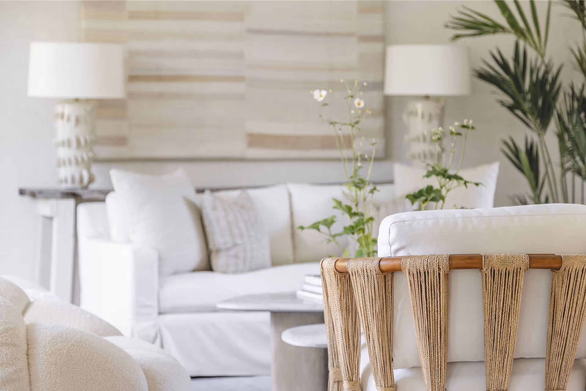 Neutral-toned living room with white sofa, textured pillows, woven-back chair, table, plants, and two white lamps.