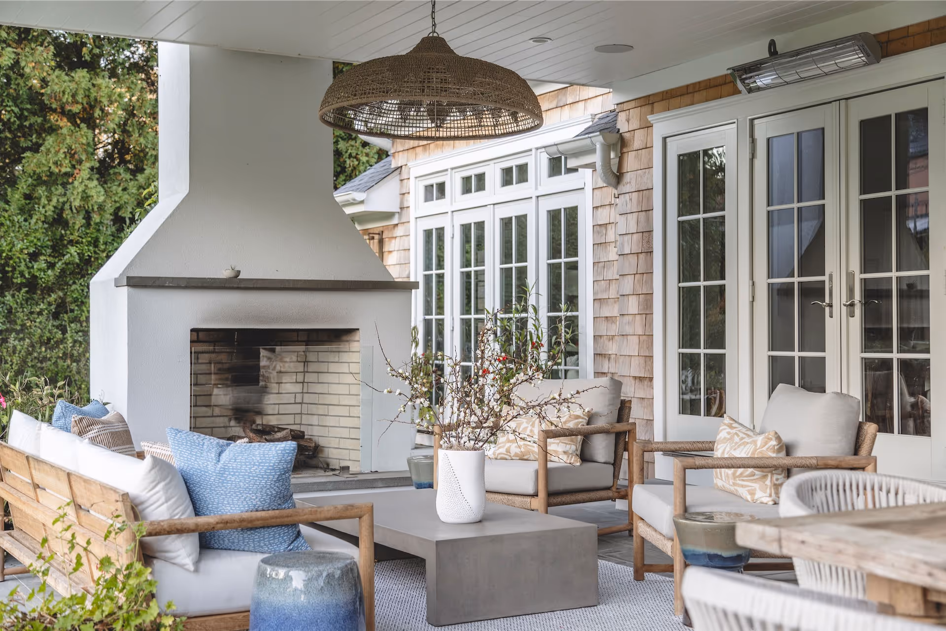 Modern outdoor patio with white fireplace, wooden furniture with cushions, concrete coffee table, and hanging wicker light fixture.