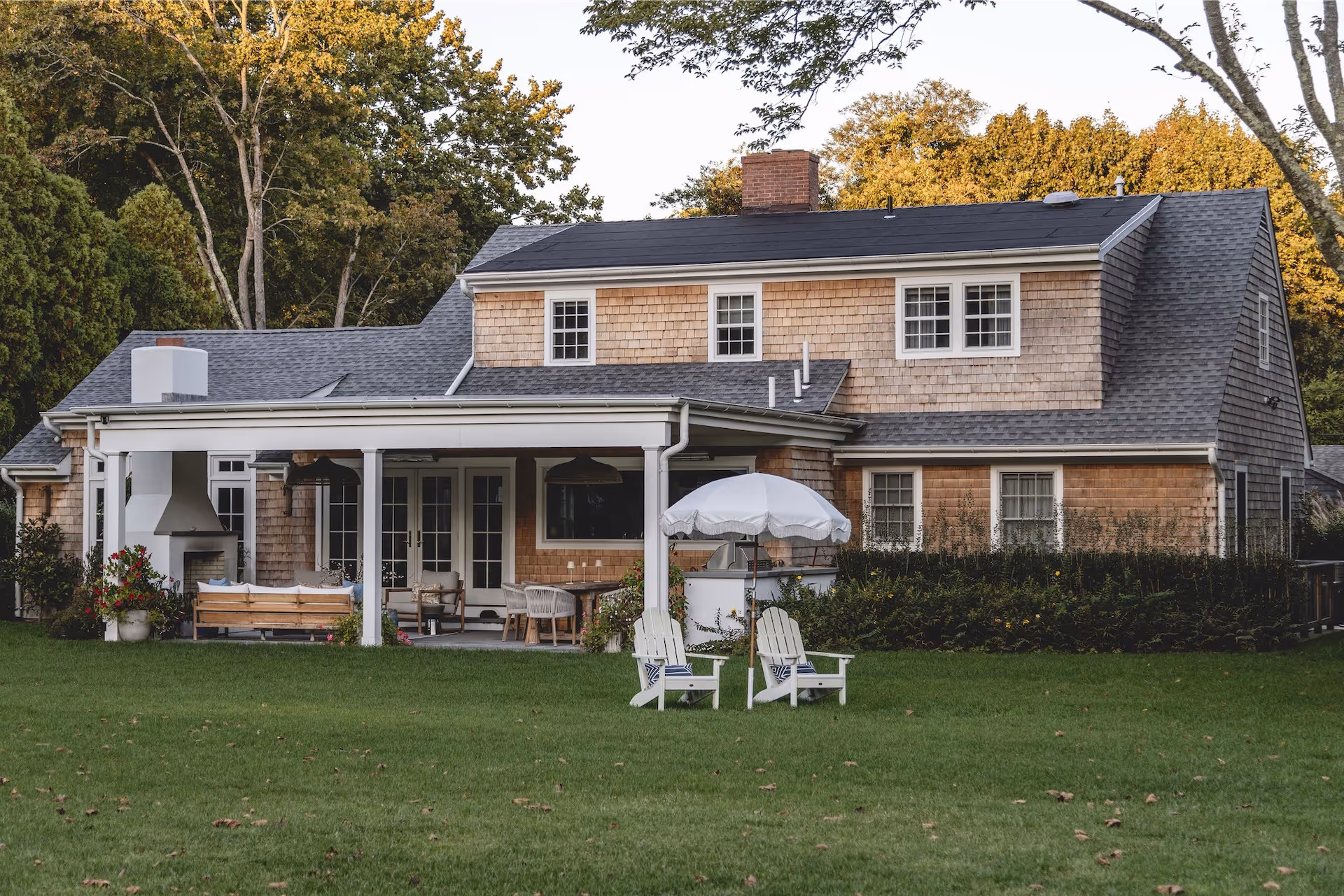 Backyard view of a cedar-shingle house with a covered patio featuring outdoor furniture, a fireplace, and two white Adirondack chairs with a white umbrella on the lawn.