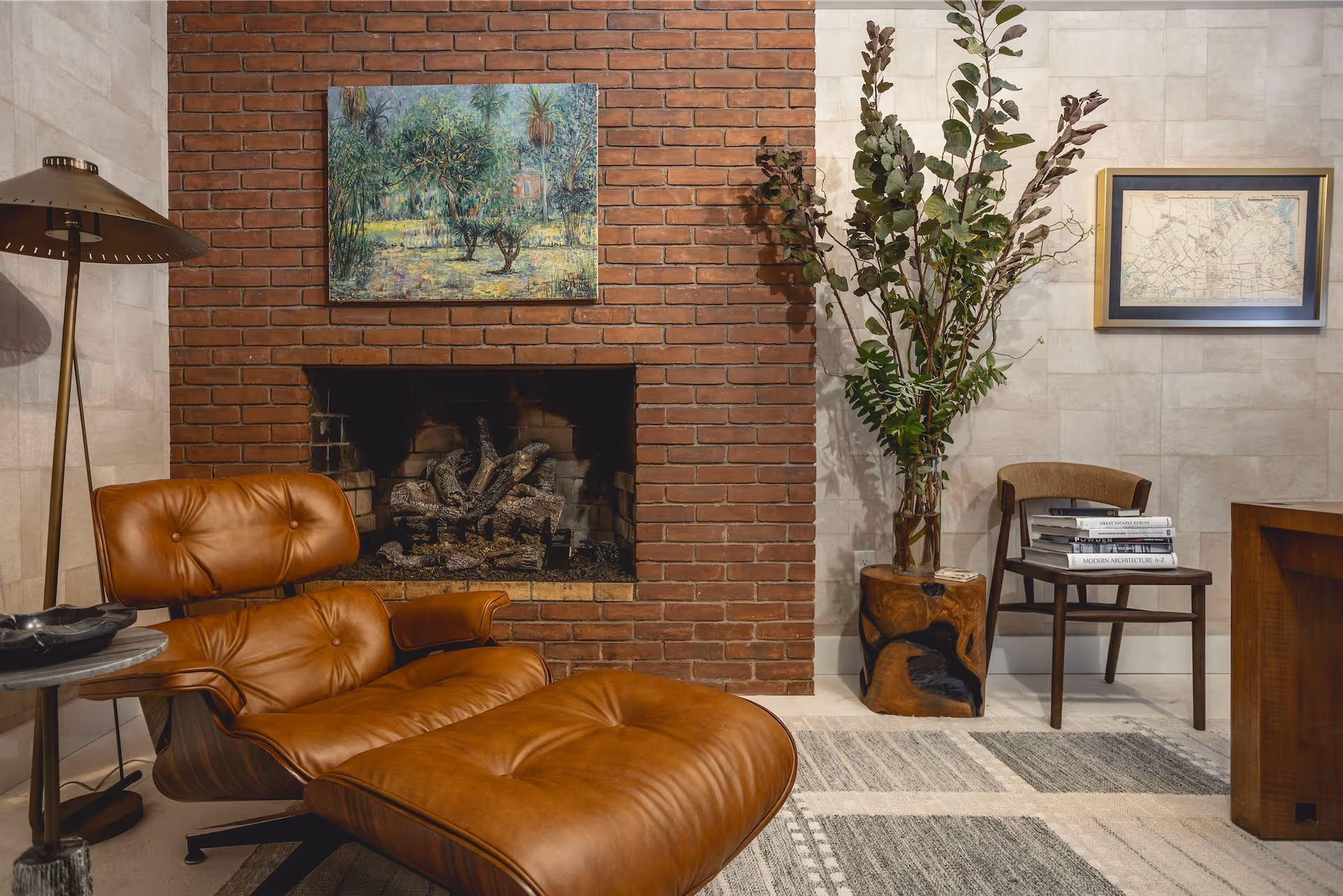 Mid-century brown leather chair with ottoman in front of a red brick fireplace, next to a floor lamp and a wooden chair with stacked books.
