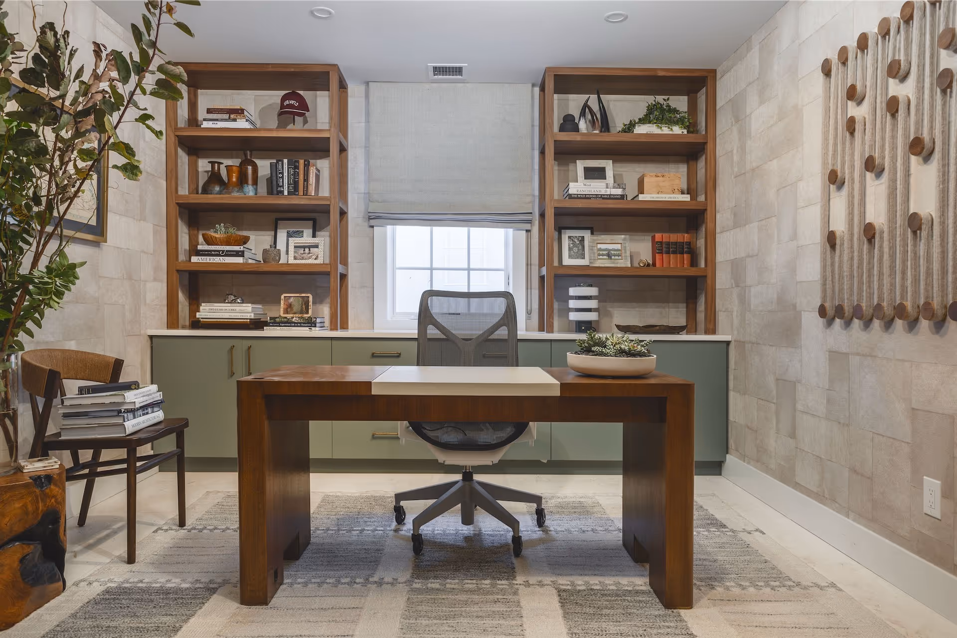 Modern home office with wooden desk, mesh office chair, built-in green cabinets, wooden shelves with books and decor, and a large plant next to a wooden chair.