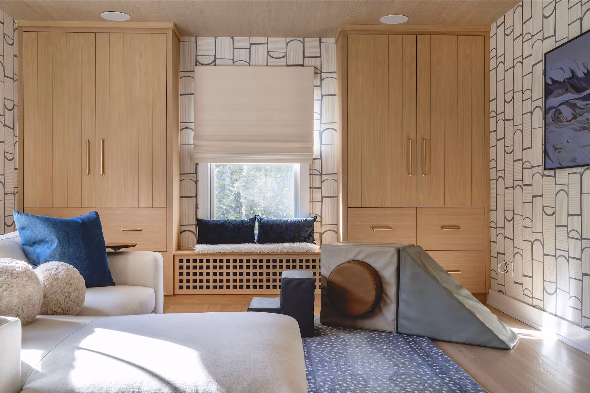 Modern playroom with light wood cabinets, window seat with blue pillows, patterned wallpaper, and soft play blocks on a blue rug.