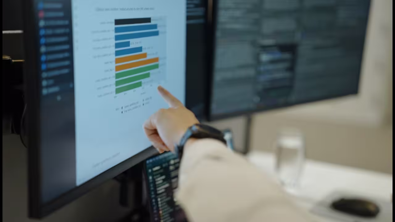 Close-up of a person pointing to performance data on a computer monitor, with code displayed on a second screen.