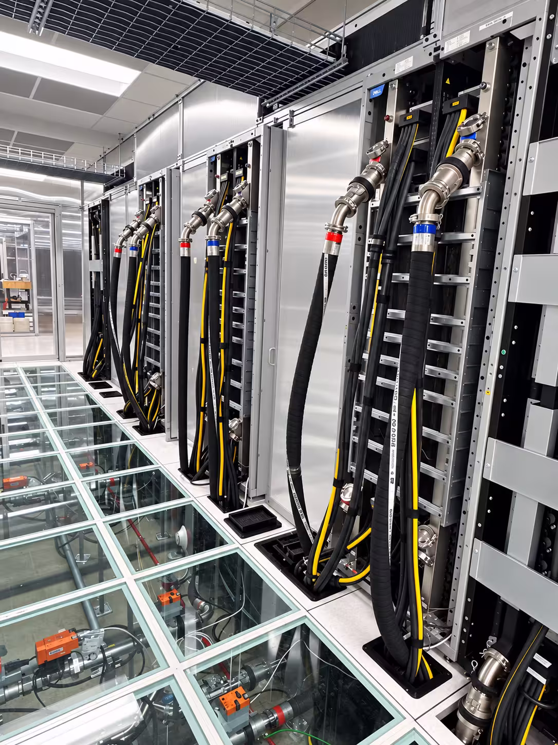 Data center aisle with a row of racks, each connected by thick black liquid cooling hoses with metal quick-disconnect fittings marked by red and blue collars. The hoses run down the face of the racks and drop through cutouts in a raised floor. Several floor tiles are transparent, showing the coolant piping, valves, and orange actuators running beneath the aisle.