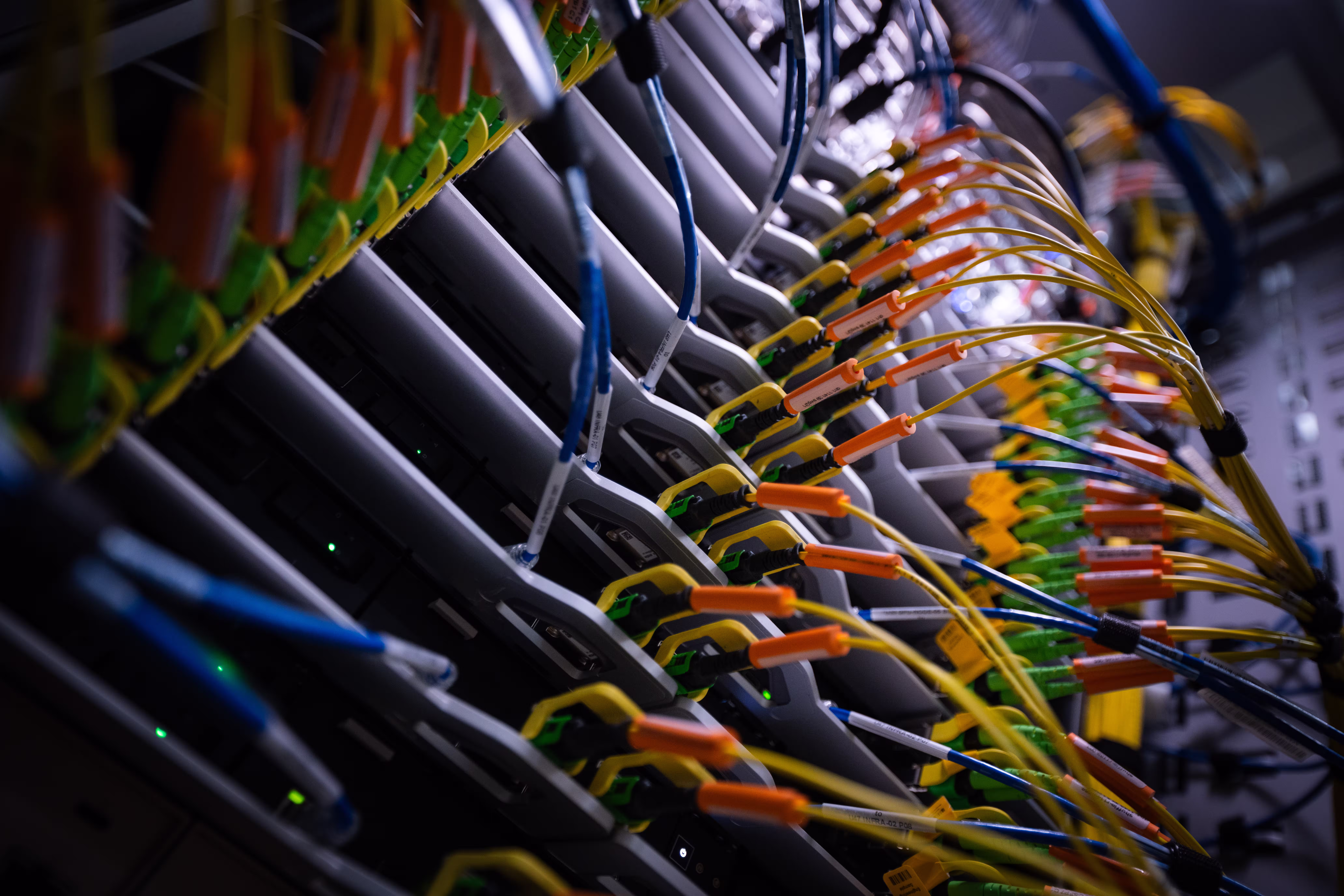 Angled close-up of networking equipment in a data center rack. Yellow single-mode fiber cables with orange and green connectors plug into labeled ports along each unit, alongside blue cables. Green status LEDs glow on the chassis; the background falls out of focus.