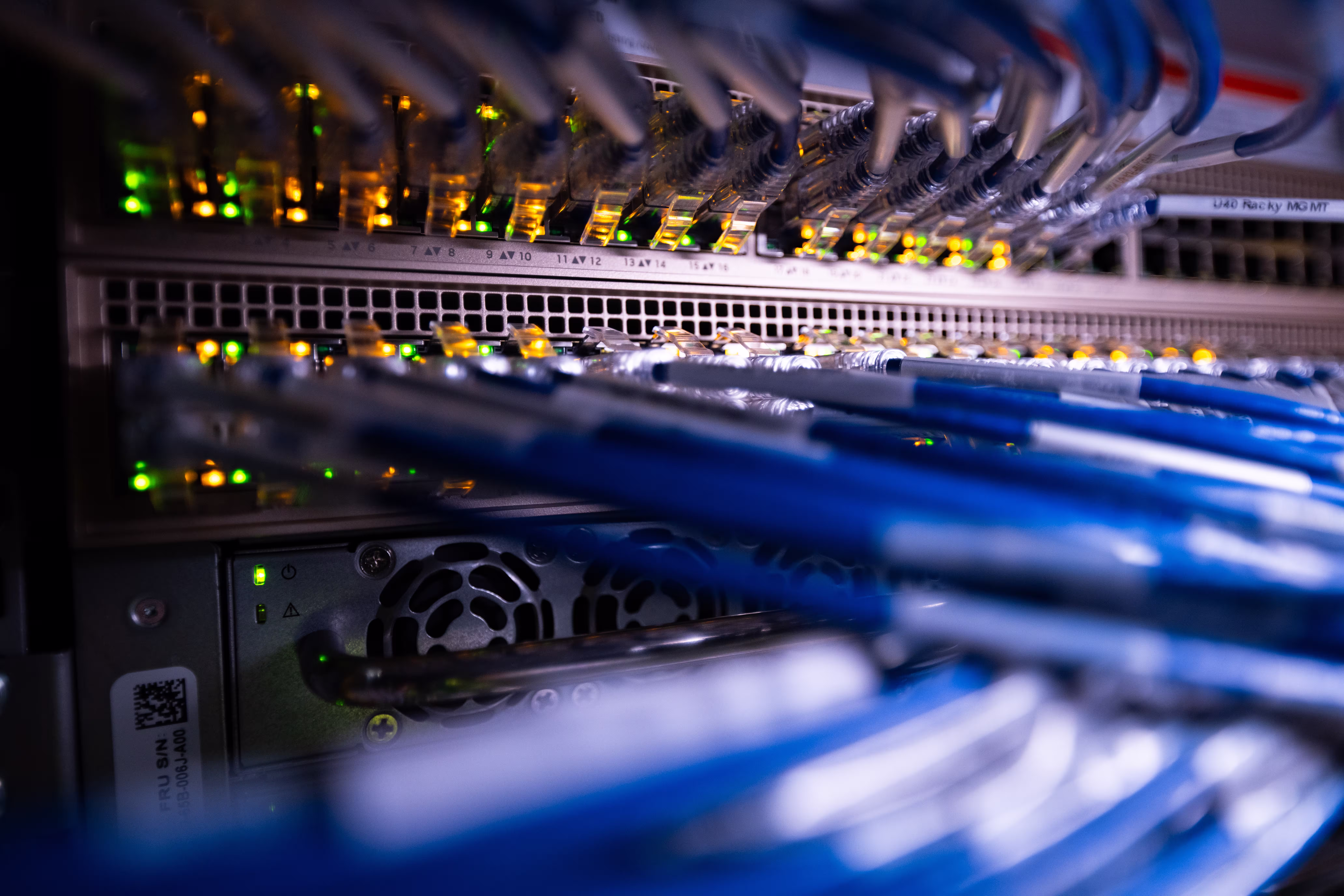Close-up of a rack switch with green and amber port LEDs lit, blue network cables running out of focus toward the camera.