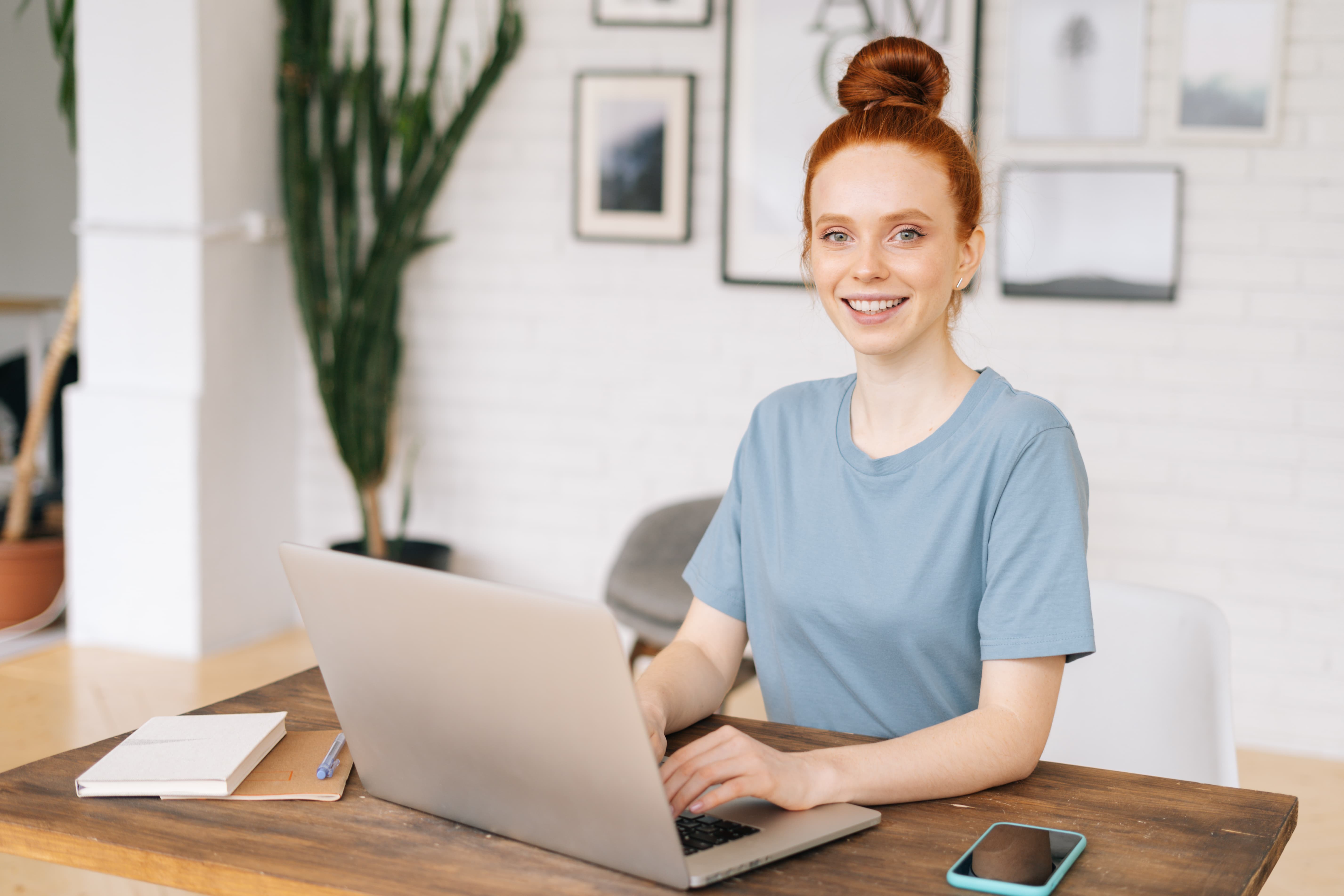 Smiling woman with long hair and wireless earbuds sitting on a wooden bench outdoors using a laptop.