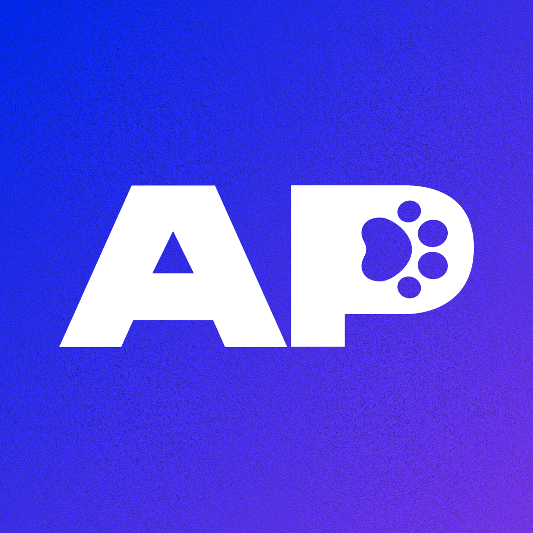 Young woman in a gray sweater writing in a notebook at a wooden kitchen table with an open laptop beside her.