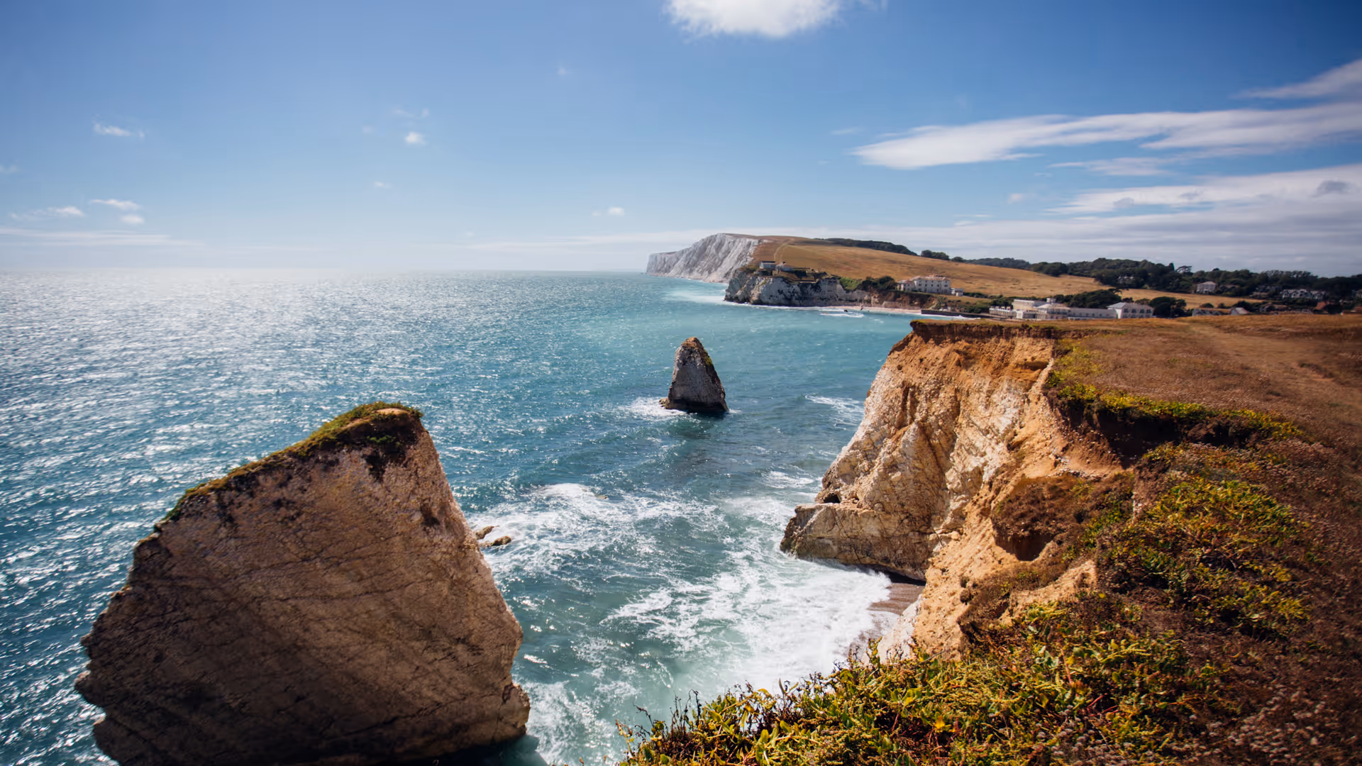 Cliffside view of a rocky coastline with turquoise sea, grassy cliffs, and a cloudy blue sky.