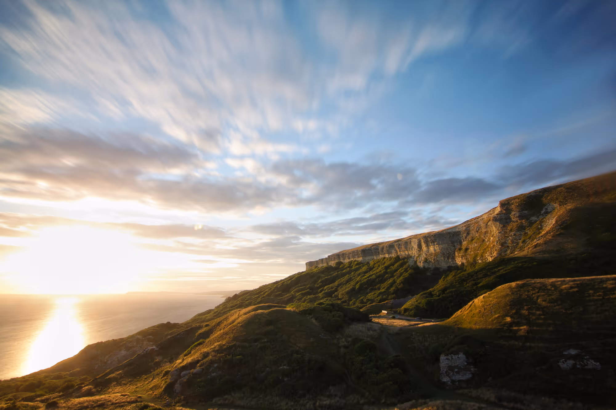 Sunset over a coastal cliff with green hills and a partly cloudy sky.