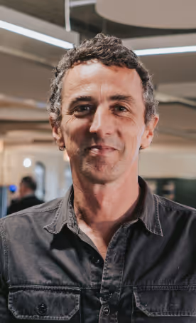Man with short curly hair and a slight smile wearing a dark button-up shirt in an indoor setting.