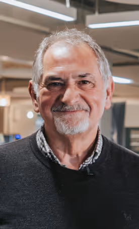 Smiling older man with gray hair and beard wearing a dark sweater and checkered shirt in an indoor setting.