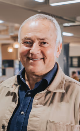 Smiling elderly man with gray hair wearing a beige jacket over a dark blue shirt indoors.