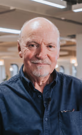 Smiling elderly man with a bald head and white beard wearing a dark blue button-up shirt in a well-lit indoor setting.
