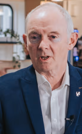 Older man in a dark blazer and white shirt speaking indoors with blurred background.