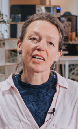 Woman with light skin and brown hair tied back, wearing a white shirt over a dark patterned top and speaking with a microphone clipped to her shirt.