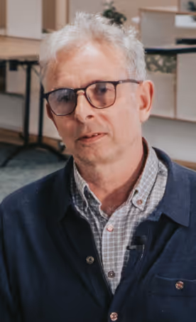 Middle-aged man with gray hair and glasses wearing a checkered shirt and dark jacket indoors.