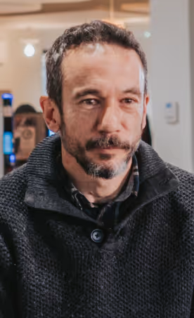 Man with short curly hair and a beard wearing a dark knitted sweater in an indoor setting.